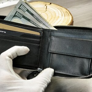 May include: Black leather wallet with a silver zip, containing several one hundred dollar bills and a credit card. The wallet is open, showing its internal compartments and contents.