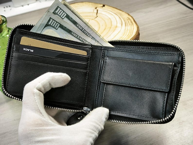 May include: Black leather wallet with a silver zip, containing several one hundred dollar bills and a credit card. The wallet is open, showing its internal compartments and contents.