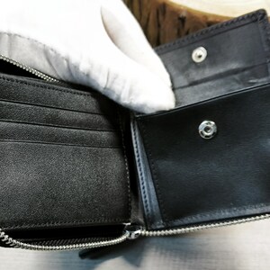 May include: Black leather wallet, open to reveal card slots and a coin pocket. The wallet has a silver zip closure and a snap fastening. The interior is black and appears to be unused.