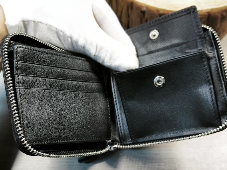 May include: Black leather wallet, open to reveal card slots and a coin pocket. The wallet has a silver zip closure and a snap fastening. The interior is black and appears to be unused.