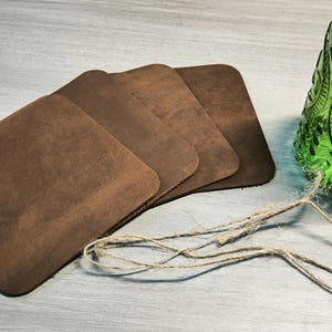 May include: Four square, brown leather coasters with rounded corners are stacked on a light gray surface. A green glass bottle with a decorative pattern and a length of twine are also in the image. The coasters are a rustic home decor item.