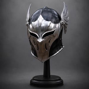 May include: A black helmet featuring a polished silver metal faceplate with sharp, angular cutouts for the eyes and mouth. The helmet is adorned with prominent, detailed silver wings attached to each side, mounted on a black circular display stand.