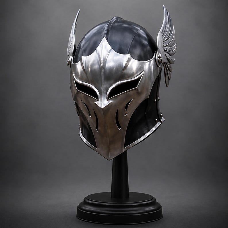 May include: A black helmet featuring a polished silver metal faceplate with sharp, angular cutouts for the eyes and mouth. The helmet is adorned with prominent, detailed silver wings attached to each side, mounted on a black circular display stand.
