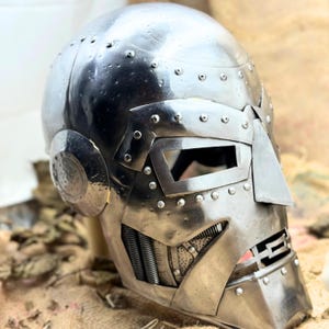May include: A silver-colored, riveted metal helmet with a distinctive angular design. The helmet features a visor with a rectangular opening, a prominent nose piece, and a detailed mouth area with mechanical elements. The overall aesthetic suggests a futuristic or sci-fi theme.