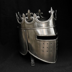 Op de afbeelding: Een zilverkleurige ridderhelm met een decoratieve kroon. De helm heeft een vizier met een rechthoekige opening en verschillende kleine ventilatiegaten. Een leren riem met een gesp is bevestigd. De achtergrond is zwart.