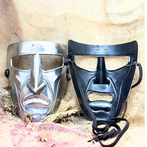 Könnte beinhalten: Zwei metallische Masken mit historischem Thema. Eine Maske ist silberfarben mit einem Netz über den Augen, die andere ist schwarz. Beide Masken haben ein markantes Nasen- und Munddesign, mit schwarzen Kordeln zur Befestigung. Die Masken werden auf einem strukturierten, neutralfarbenen Hintergrund präsentiert.