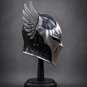May include: A black metal helmet featuring a polished silver faceplate with narrow eye slits and a prominent silver wing attachment on the side. The helmet is constructed with visible rivets along the side and is displayed on a black stand.