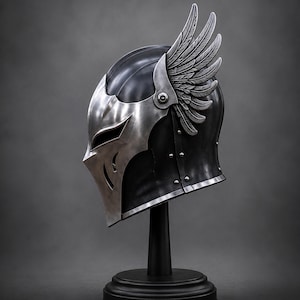 May include: A metallic helmet featuring a dark, smooth finish and a silver-toned faceplate with angular cutouts. The helmet is adorned with a prominent, detailed silver wing attachment on the side, secured with a visible rivet, and a silver trim.