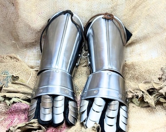 May include: Pair of polished steel gauntlets, featuring articulated finger plates and protective cuffs. The gauntlets are designed with a medieval aesthetic, showcasing riveted construction and a smooth, reflective surface. The gloves are displayed on a textured, neutral-toned background.