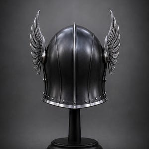 May include: A dark gray metal helmet featuring a central vertical ridge and two prominent, upward-curving silver wings attached to the sides. The helmet has a smooth, rounded dome, a flared base with visible rivets, and a polished silver trim edge.