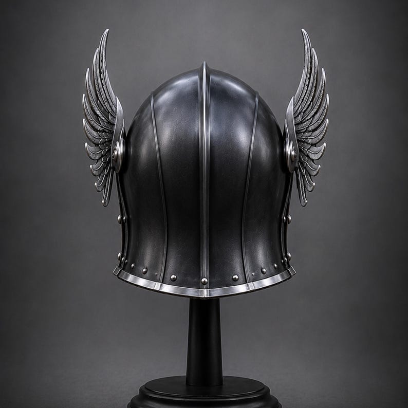 May include: A dark gray metal helmet featuring a central vertical ridge and two prominent, upward-curving silver wings attached to the sides. The helmet has a smooth, rounded dome, a flared base with visible rivets, and a polished silver trim edge.