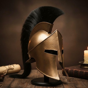 Op de afbeelding: Een bronzen Spartaanse helm met een zwarte pluim, tentoongesteld op een zwarte standaard. De helm heeft een klassiek ontwerp met een prominente wenkbrauw en wangbeschermers. Een opgerold perkament en een kaars staan op de achtergrond.