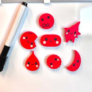 Può includere: Una collezione di calamite rosse in stile cartone animato in varie forme, tra cui cerchi, una goccia, una luna crescente e una stella. Ogni calamita presenta un volto sorridente con occhi neri e dettagli bianchi. Sono visibili anche una lavagna e un pennarello.