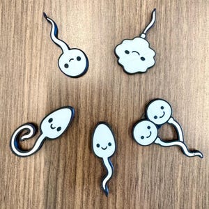 May include: Five white sperm-shaped cutouts with black outlines and smiling faces are arranged on a wooden surface. Each cutout has a unique shape, with varying tail designs. The sperm shapes are a light blue colour.