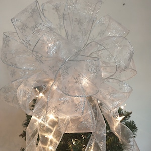 Peut inclure: Un grand nœud blanc pour sapin de Noël avec des motifs de flocons de neige argentés. Le nœud est fait de ruban transparent et est illuminé par de petites lumières blanches, créant une décoration de vacances festive.