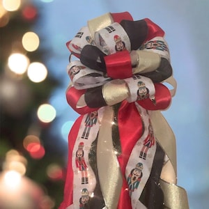 May include: A festive Christmas tree topper bow featuring red, black, gold, and white ribbons. The white ribbon is printed with a nutcracker soldier design. The bow is adorned with small, twinkling lights, creating a holiday atmosphere.
