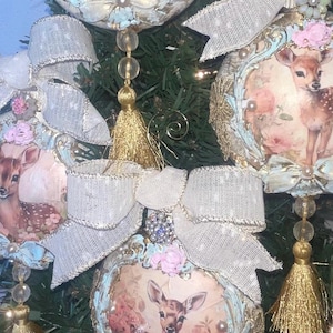 May include: Three round Christmas ornaments featuring a fawn illustration surrounded by pink floral accents. Each ornament is topped with a large, textured white ribbon bow and includes a dangling gold tassel and clear beads at the bottom.