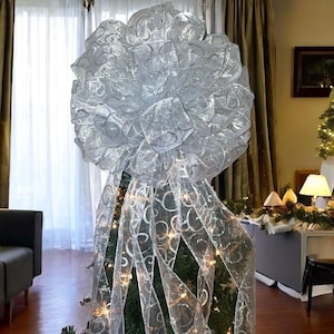 May include: A large, silver Christmas tree bow made of sheer, patterned ribbon. The bow sits atop a decorated evergreen tree with twinkling lights. The ribbon cascades down the tree, creating a festive holiday display.