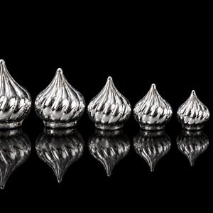 May include: A set of six silver-coloured, metallic, onion-shaped decorative finials arranged in a row from largest to smallest. Each piece features a spiralling, ribbed texture that tapers to a point at the top and has a flat base.