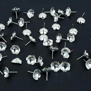 May include: A selection of small, silver-coloured flower-shaped jewellery components scattered on a dark surface. Each component features a petal design and a post for attaching to other jewellery pieces. The components are likely for earrings or other small jewellery.