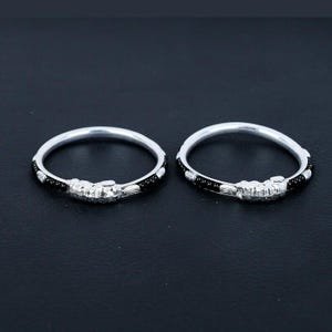 May include: Two silver bangles with intricate designs. Each bangle features a central decorative element and is accented with black beads. The bangles are displayed on a dark surface, showcasing their reflective qualities.