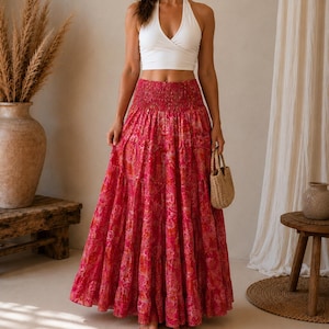 Puede incluir: Un top corto blanco estilo halter con diseño cruzado al frente, combinado con una falda larga maxi de niveles en un vibrante estampado floral tipo cachemira en tonos rosa y naranja, con cintura elástica fruncida y silueta amplia.