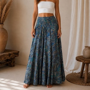 Pode incluir: Um top cai-cai branco sem alças combinado com uma saia comprida até ao chão com um padrão paisley em tons de azul, verde-azulado e dourado. A saia tem uma cintura elástica larga e franzida e uma silhueta ampla e fluida.