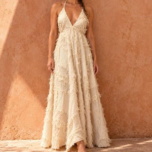 May include: A floor-length, cream-colored halter dress featuring a patchwork design with frayed edges. The garment has a deep V-neckline, thin straps that tie at the neck, and a flowing, loose-fitting skirt constructed from textured fabric panels.