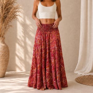 May include: A floor-length maxi skirt featuring a vibrant red and orange paisley pattern with a wide, gathered smocked waistband. The skirt has a full, flowing silhouette that reaches the floor, crafted from a lightweight fabric with a detailed print.