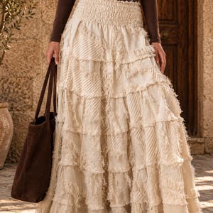 May include: A floor-length, off-white tiered skirt featuring a smocked elastic waistband and raw, frayed edges on each horizontal panel. The fabric displays a mix of textures, including solid sections and subtle diagonal woven patterns throughout.