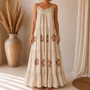 Könnte beinhalten: Ein bodenlanges, gestuftes Kleid in Beige mit dünnen Spaghettiträgern, einem gehäkelten Oberteil und einem sich wiederholenden braunen Blumenmuster auf dem Rock. Das Kleidungsstück hat eine lockere, fließende Silhouette mit gerafften Stofflagen.