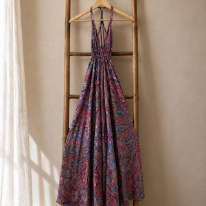Puede incluir: Un vestido largo tipo halter con un vibrante estampado de cachemira en tonos magenta, morado, azul y dorado. La prenda presenta un escote en V profundo, tirantes finos que se cruzan en la espalda y una cintura fruncida con falda amplia.