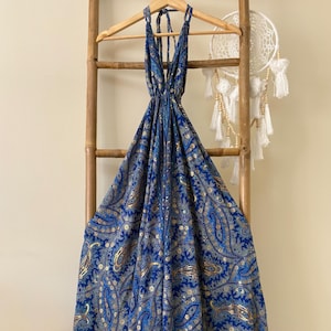 May include: A blue paisley print dress with a halter neckline hangs on a wooden hanger. The dress features a vibrant pattern in shades of blue and gold. The dress is displayed on a bamboo ladder, with a dreamcatcher in the background.