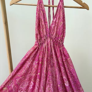May include: A bright pink halter dress with a plunging neckline and a flowing skirt. The dress has a paisley pattern in pink and gold. It is displayed on a wooden hanger, highlighting its design and style.