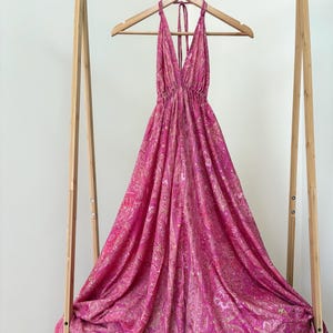 May include: A vibrant, halter-neck dress in pink and magenta hues, featuring a detailed paisley design. The dress is displayed on a wooden hanger, revealing its full length and pattern. The fabric flows gracefully, emphasizing the dress's lightweight quality.