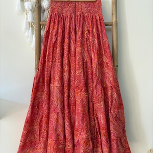 May include: A long, flowing skirt in shades of red, pink, and orange, with a detailed paisley pattern. The skirt has a smocked waistband and hangs from a wooden hanger. The fabric appears lightweight. The skirt's length is suitable for warm weather.