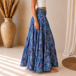 Op de afbeelding: Een enkellange, gelaagde rok in blauw met een gedetailleerd paisley-patroon in marineblauw, lichtblauw en goud. De rok heeft een brede, gesmokte elastische tailleband en een wijd uitlopend, vloeiend silhouet dat tot op de grond reikt.