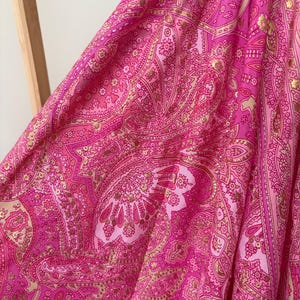 May include: Close-up of a vibrant pink fabric with a detailed paisley pattern. The design incorporates gold accents, creating a luxurious textile. The fabric is draped, showcasing its flow and texture.