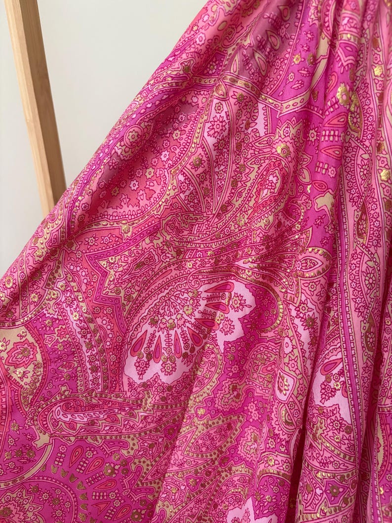 May include: Close-up of a vibrant pink fabric with a detailed paisley pattern. The design incorporates gold accents, creating a luxurious textile. The fabric is draped, showcasing its flow and texture.