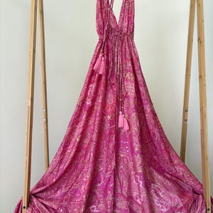 May include: A bright pink halter dress with a paisley design and tassel details. The dress is displayed on a wooden hanger and stand. The fabric appears lightweight and flowing, ideal for warmer climates. The dress is approximately 150 cm in length.