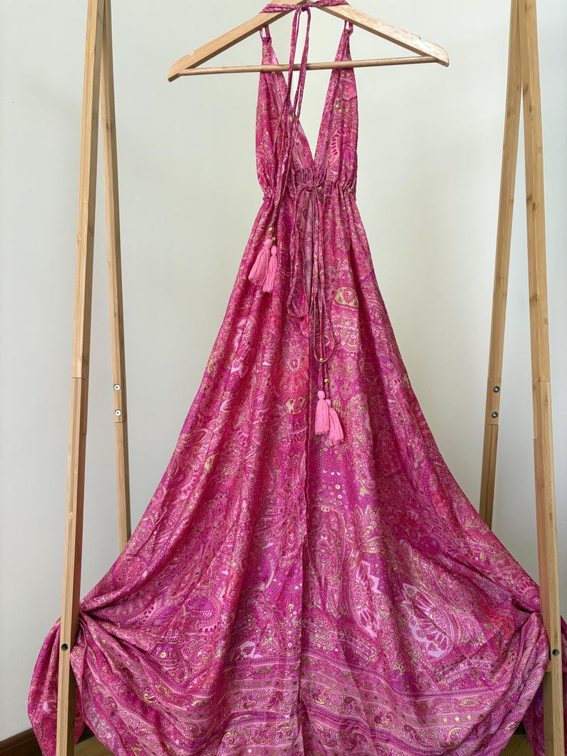 May include: A bright pink halter dress with a paisley design and tassel details. The dress is displayed on a wooden hanger and stand. The fabric appears lightweight and flowing, ideal for warmer climates. The dress is approximately 150 cm in length.