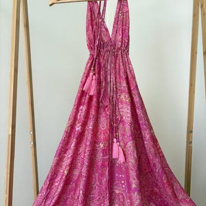 May include: A bright pink halter dress with a paisley design and a deep V-neck. The dress has a drawstring at the waist and pink tassels. It is displayed on a wooden hanger.