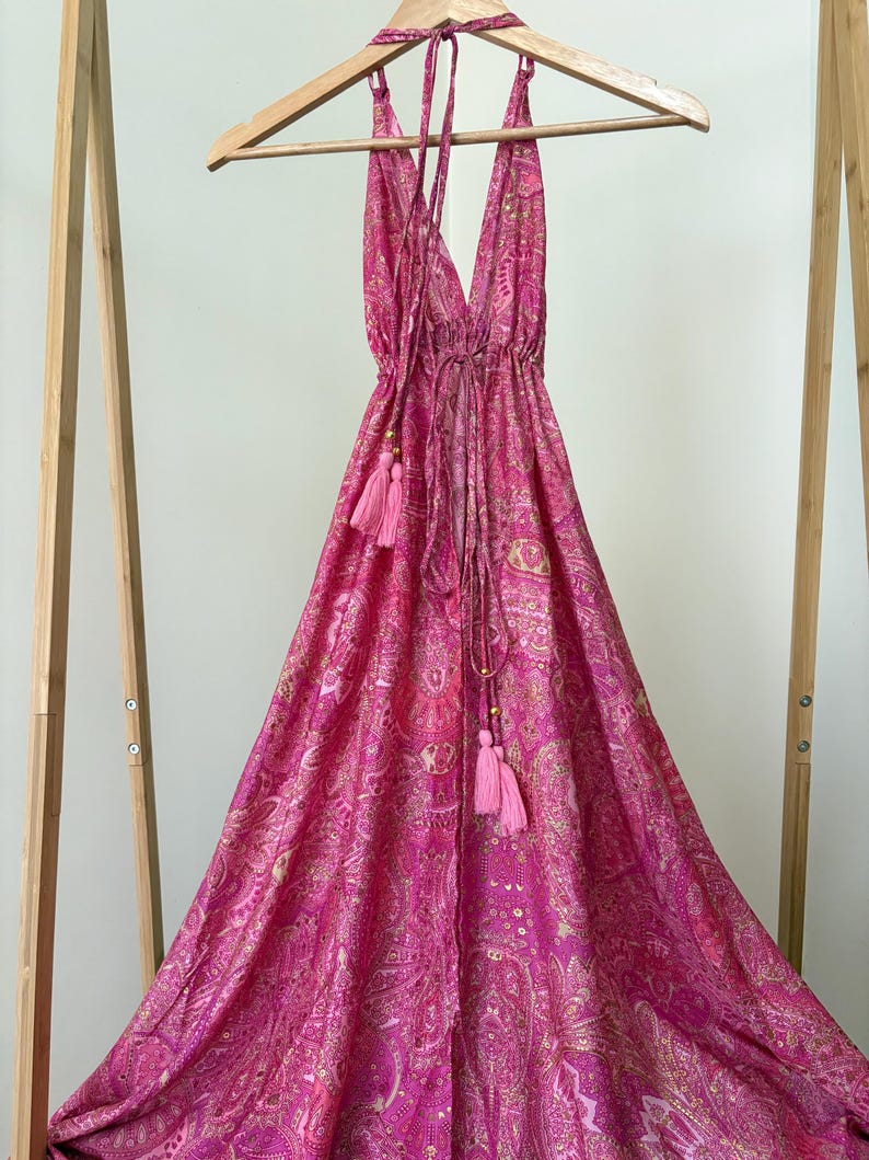 May include: A bright pink halter dress with a paisley design and a deep V-neck. The dress has a drawstring at the waist and pink tassels. It is displayed on a wooden hanger.