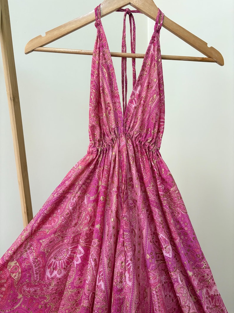 May include: A bright pink halter dress with a plunging neckline and a flowing skirt. The dress has a paisley pattern in pink and gold. It is displayed on a wooden hanger, highlighting its design and style.