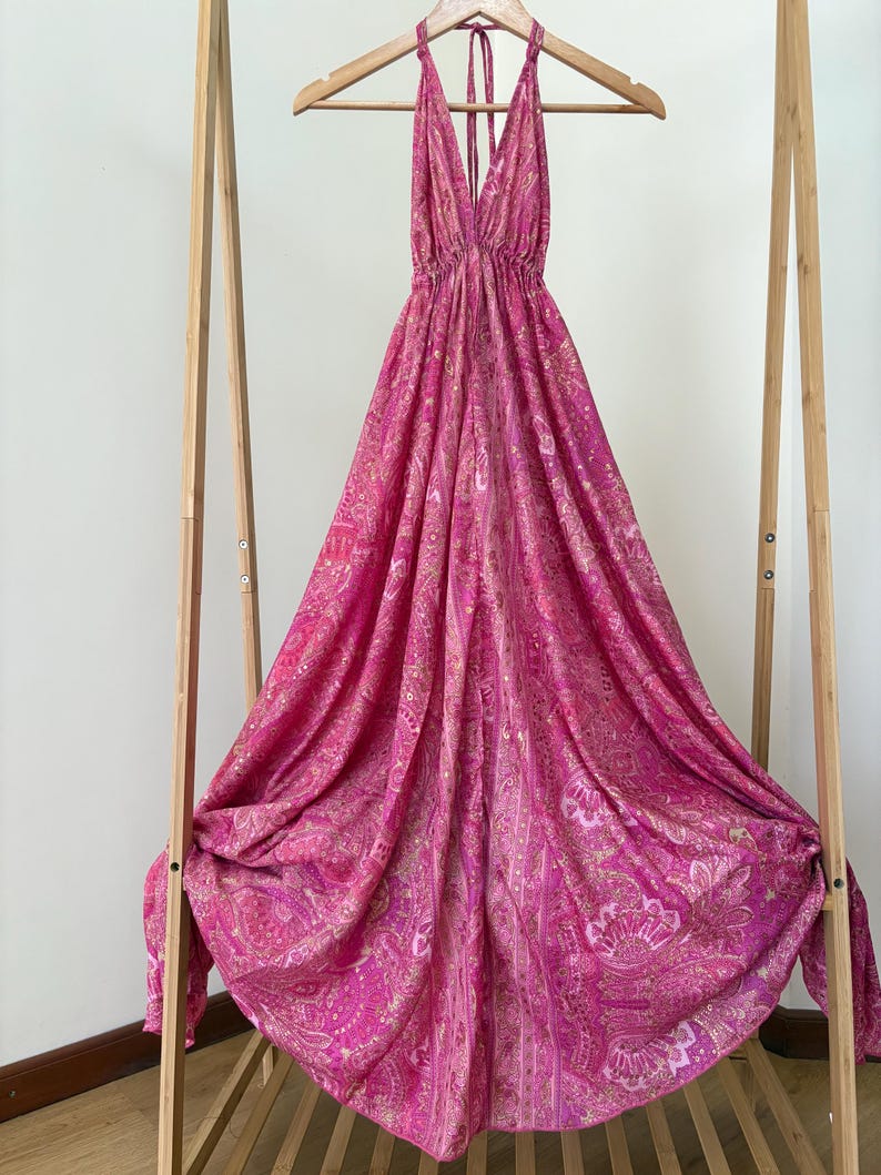 May include: A vibrant, halter-neck dress in pink and magenta hues, featuring a detailed paisley design. The dress is displayed on a wooden hanger, revealing its full length and pattern. The fabric flows gracefully, emphasizing the dress's lightweight quality.