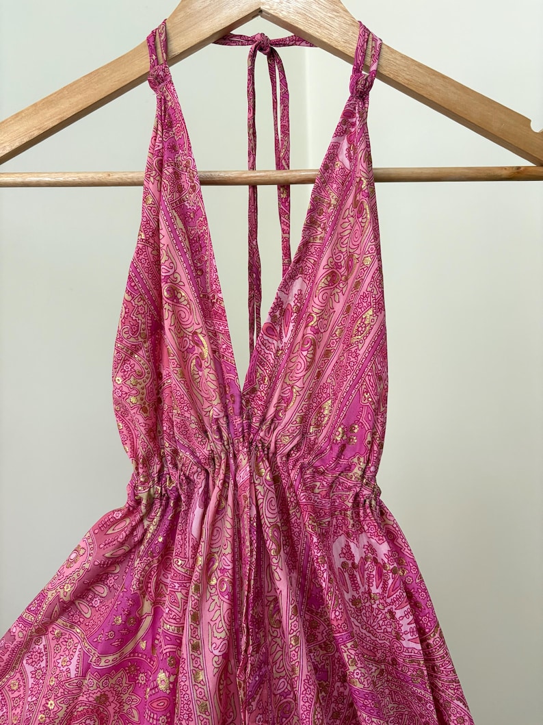 May include: A pink halter top with a deep V-neck and a paisley pattern. The top is hanging on a wooden hanger. The fabric has a flowing, lightweight appearance.
