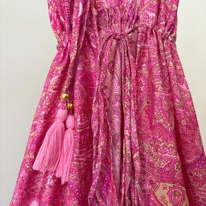 May include: A pink paisley patterned dress with a deep V-neck and a drawstring waist. The dress features pink tassels with gold beads. The fabric has a shiny appearance.
