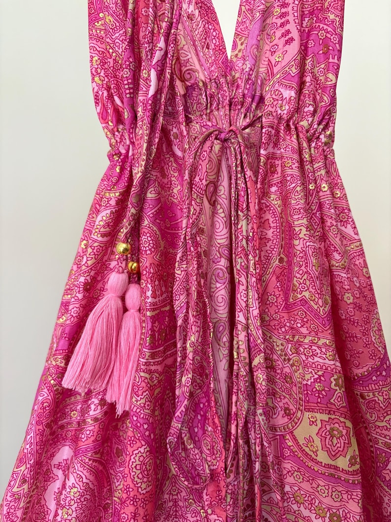 May include: A pink paisley patterned dress with a deep V-neck and a drawstring waist. The dress features pink tassels with gold beads. The fabric has a shiny appearance.