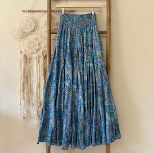 Può includere: Una gonna lunga e fluida con un motivo paisley blu, appesa a una scala di legno. La gonna ha un punto vita arricciato e un design a più livelli. Il tessuto presenta sfumature di blu, verde acqua e oro. La gonna è lunga circa 91 cm.