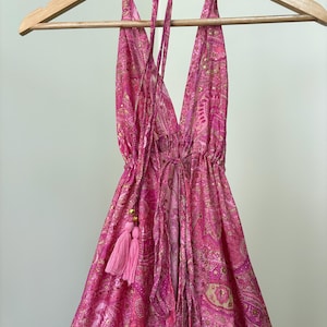 May include: A pink paisley halter dress hanging on a wooden clothes hanger. The dress has a deep V-neck and drawstring waist. A pink tassel hangs from the side. The dress is made of a lightweight fabric.